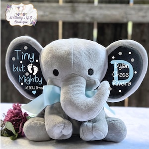 May include: A gray plush elephant with a light blue bow, featuring the text "Tiny but Mighty" and "NICU Grad" on one ear, and the name "Dylan Chase Rivera" on the other. The elephant has black eyes and is surrounded by flowers.