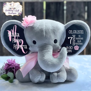 May include: A gray plush elephant with a pink bow, flower, and personalized text on the ears. The text includes a name, date, weight (7 pounds, 11 ounces), and length (19.5 inches). The elephant has black eyes and a pink flower on its head.