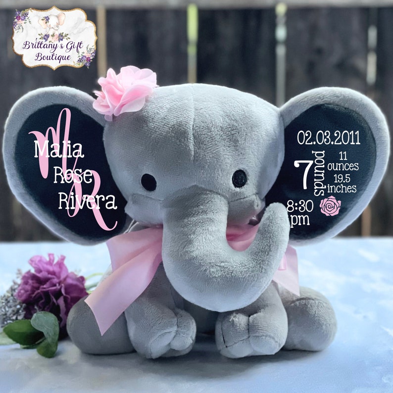 May include: A gray plush elephant with a pink bow, flower, and personalized text on the ears. The text includes a name, date, weight (7 pounds, 11 ounces), and length (19.5 inches). The elephant has black eyes and a pink flower on its head.