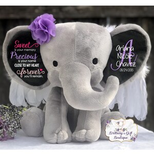 May include: A gray plush elephant with a purple flower on its head and white feather accents. The elephant's ears have personalized text: "Sweet is your memory, Precious is your name, Close to my heart forever you'll remain." The other ear has a name and date.