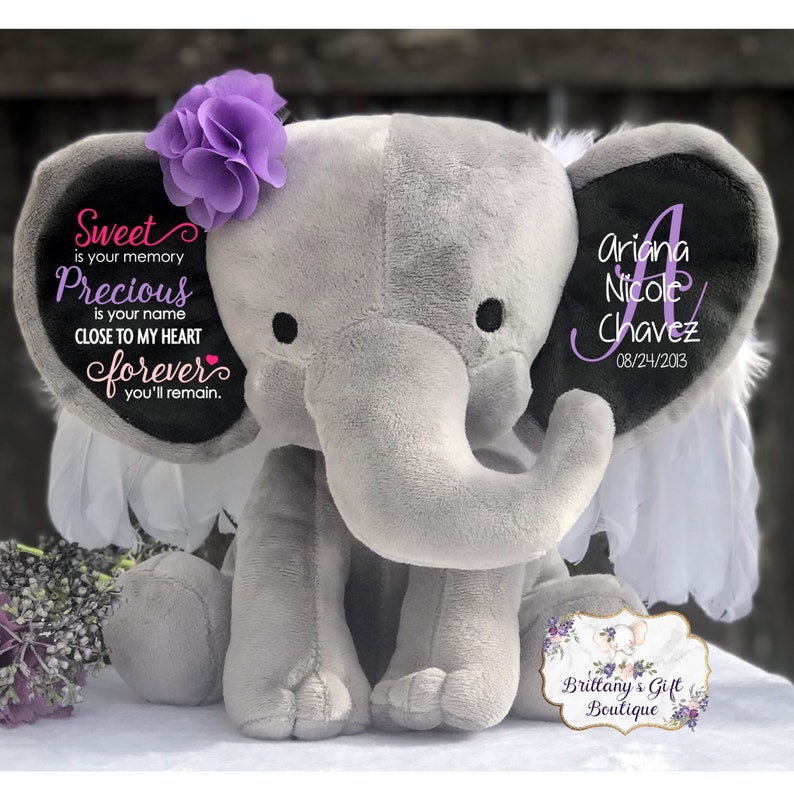 May include: A gray plush elephant with a purple flower on its head and white feather accents. The elephant's ears have personalized text: "Sweet is your memory, Precious is your name, Close to my heart forever you'll remain." The other ear has a name and date.