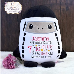 May include: A plush, white and black astronaut-shaped pillow with embroidered details. The pillow features a smiling face and the name "Jasmine", birth details: 7 lbs 10 oz, 21 1/2 inches, and the date March 19, 2025.