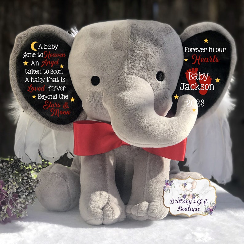 Puede incluir: Un elefante de peluche gris con una pajarita roja y alas. El elefante tiene texto bordado en sus orejas: "A baby gone to Heaven An Angel taken to soon A baby that is Loved forever Beyond the Stars & Moon" y "Forever in our Hearts Baby Jackson 2023".