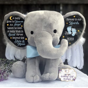 Puede incluir: Un elefante de peluche gris con alas azules y un lazo azul. El elefante tiene un parche de fieltro negro en cada oreja con texto blanco. La oreja izquierda dice "A baby gone to Heaven ★ An Angel ★ taken to soon A baby that is Loved forver ★ Beyond the Stars & Moon". La oreja derecha dice "Forever in our ★ Hearts ★ Baby Jackson ★ 2023".