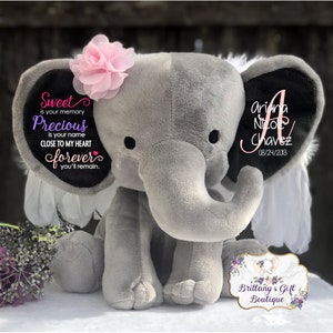May include: A gray plush elephant with black ears and a pink flower on its head. Text on the elephant includes the name "Ariana Nicole Chavez" and the date "08/24/2013". Additional text reads "Sweet is your memory, Precious is your name, Close to my heart forever you'll remain."