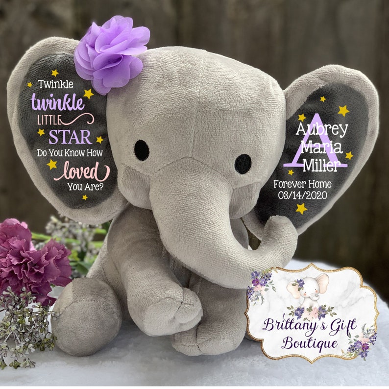 May include: A plush gray elephant toy with a purple fabric flower on its left ear. The left ear features the text Twinkle twinkle LITTLE STAR Do You Know How loved You Are? with yellow stars. The right ear displays the name Aubrey Maria Miller.