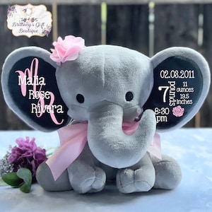 Puede incluir: Un elefante de peluche gris con un lazo y una flor rosas, con texto personalizado en las orejas. El texto incluye un nombre, fecha, peso (3,18 kg) y longitud (49,5 cm). Una flor rosa adorna la cabeza del elefante.