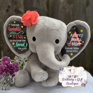 May include: A plush gray elephant toy with a coral fabric flower on its head. The left ear features the text Twinkle Twinkle Little Star Do You Know How Loved You Are? with yellow stars. The right ear displays the name Aubrey Maria Miller.