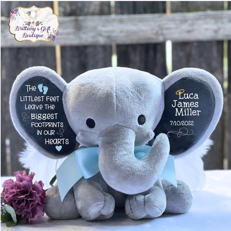 Op de afbeelding: Een grijze pluche olifant met blauwe vleugels en een zwart vilten hart op zijn borst. De oren van de olifant zijn versierd met zwart vilt en witte tekst die "The littlest feet leave the biggest footprints in our hearts" en "Luca James Miller 7/10/2022" luidt.