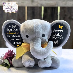 Puede incluir: Un elefante de peluche gris con un lazo amarillo y ojos negros. Las orejas del elefante tienen el texto "A hug from heaven for when you need it most" y "Sweet Baby Marks". Un regalo para beb&eacute;s.