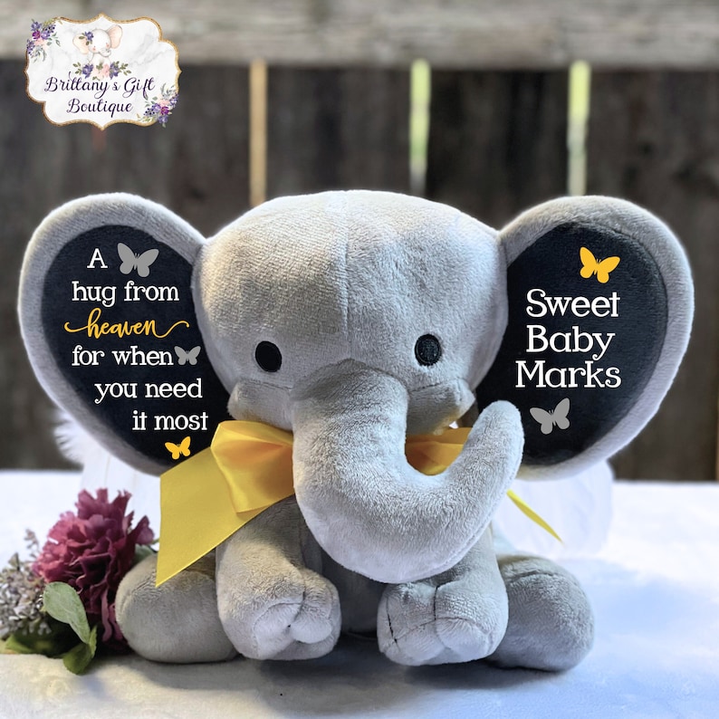 Puede incluir: Un elefante de peluche gris con un lazo amarillo y ojos negros. Las orejas del elefante tienen el texto "A hug from heaven for when you need it most" y "Sweet Baby Marks". Un regalo para beb&eacute;s.