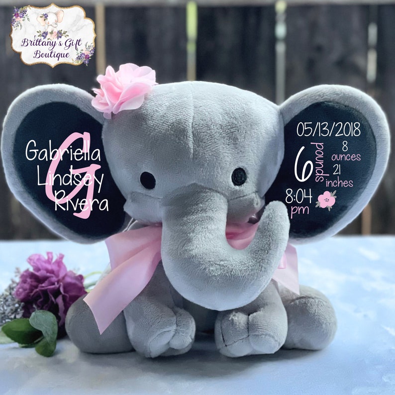 K&ouml;nnte beinhalten: Ein graues Pl&uuml;schelefant-Spielzeug mit einer rosa Stoffblume auf dem Kopf und einem rosa Band um den Hals. Die Ohren sind mit Text bedruckt, darunter der Name Gabriella Lindsey Rivera, das Datum 05/13/2018, 6 pounds 8 ounces und 21 inches.