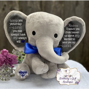 Puede incluir: Un elefante de peluche gris con un lazo azul, bordado con el texto "Loved you yesterday, Love you still, always have and always will" en una oreja y "Andrew, I watch you from heaven up above and wanted to send you my love" en la otra oreja. El elefante est&aacute; sentado sobre una superficie blanca con una peque&ntilde;a etiqueta que dice "From Grandpa Bob" y una etiqueta m&aacute;s grande que dice "Brittany's Gift Boutique".