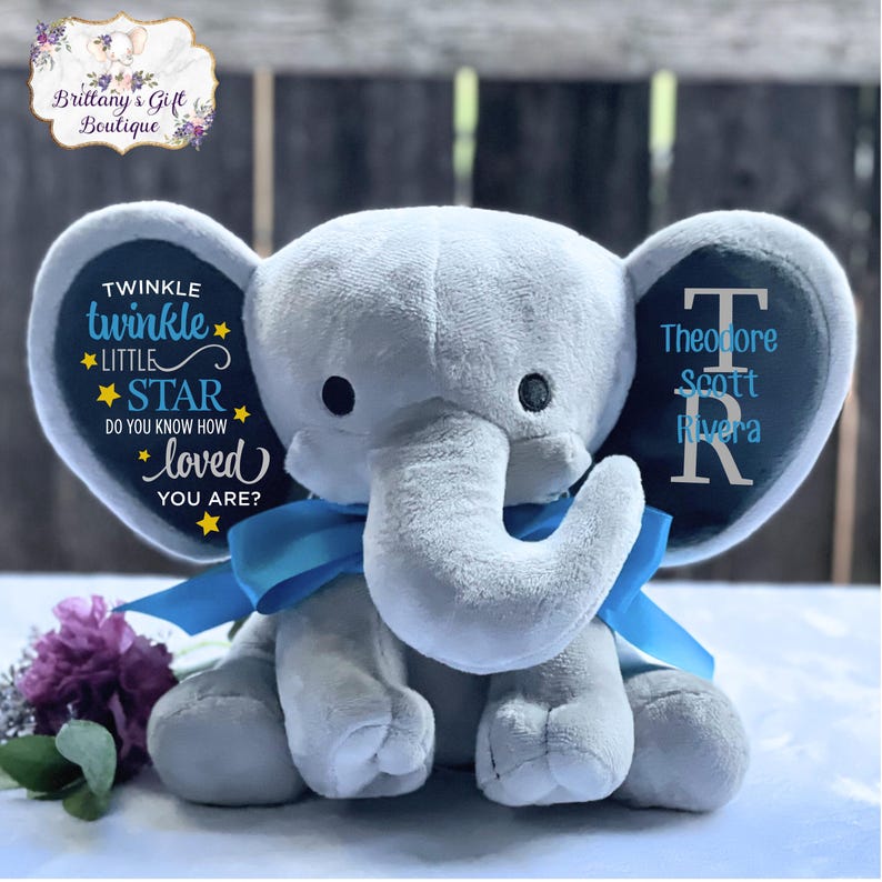 Puede incluir: Un elefante de peluche gris con detalles azules, incluyendo un lazo y detalles en las orejas. Una oreja presenta el texto "Twinkle Twinkle Little Star", la otra un nombre y una inicial. Un ramo de flores moradas est&aacute; en primer plano.
