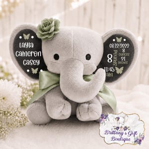 May include: A plush grey elephant toy with a sage green ribbon and a matching fabric flower on its head. The ears feature white text: Layla Cameron Casey on the left, and birth details including 08/12/2022, 8 pounds, 21 inches, 11:45 am on the right.
