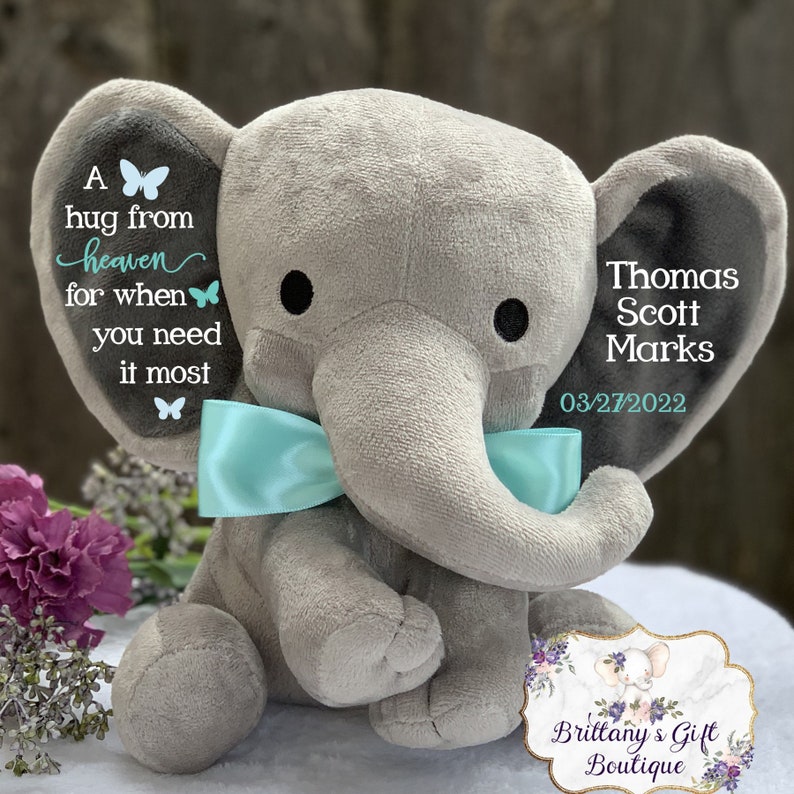 Puede incluir: Un peluche de elefante gris con una pajarita azul claro. El elefante tiene ojos negros y texto en sus orejas que dice "A hug from heaven for when you need it most" y "Thomas Scott Marks 03/27/2022".