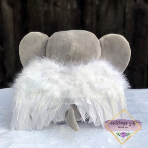 May include: A gray plush elephant with large ears and a small tail, wearing white feather angel wings. The wings are attached to the elephant's back. The image includes a logo that says "brittany's gift boutique".