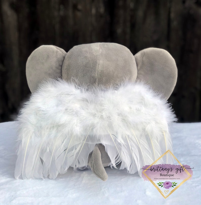 May include: A gray plush elephant with large ears and a small tail, wearing white feather angel wings. The wings are attached to the elephant's back. The image includes a logo that says "brittany's gift boutique".