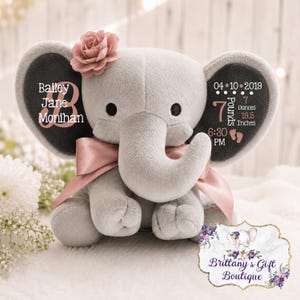 May include: A grey plush elephant toy with a pink satin ribbon around its neck and a pink fabric flower on its left ear. The left ear displays the name Bailey Jane Monihan, while the right ear shows birth details: 04-10-2019, 7 Pounds, 19.5 Inches, 6:30 PM.