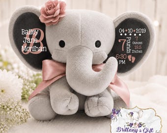 May include: A grey plush elephant toy with a pink satin ribbon around its neck and a pink fabric flower on its left ear. The left ear displays the name Bailey Jane Monihan, while the right ear shows birth details: 04-10-2019, 7 Pounds, 19.5 Inches, 6:30 PM.