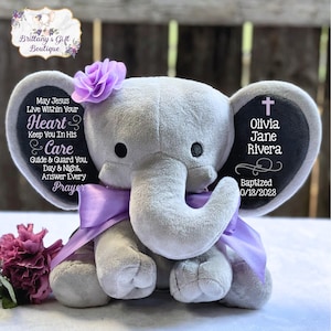 May include: A gray plush elephant toy with a purple bow and a flower on its head. The elephant has a black ear with the text "Olivia Jane Rivera Baptized 10/13/2023" and a cross. The other ear has a message about Jesus living within your heart and care.