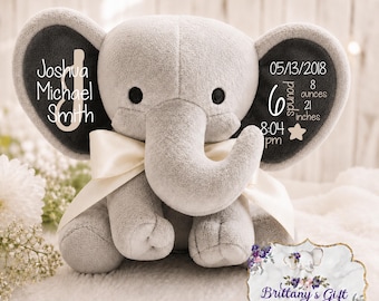 May include: A plush gray elephant toy with a cream-colored satin ribbon tied around its neck. The elephant's ears feature white text: Joshua Michael Smith on the left, and 05/13/2018, 6 pounds 8 ounces, 21 inches, and 8:04 pm on the right.