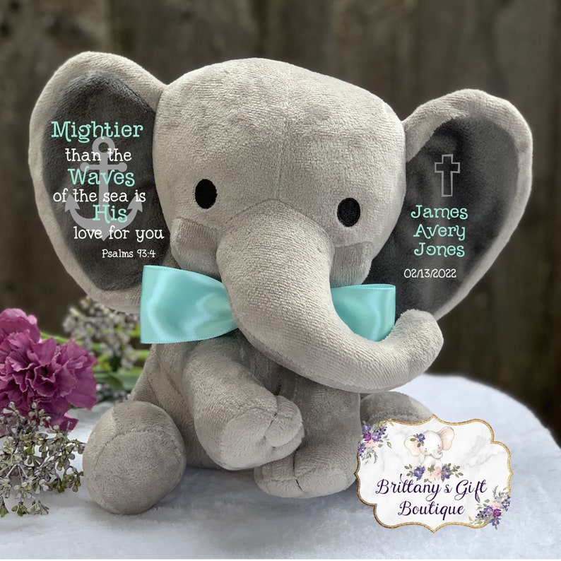 May include: A plush gray elephant toy featuring a light blue satin bow around its neck. The left ear displays an anchor icon and the text "Mightier than the Waves of the sea is His love for you Psalms 93:4". The right ear shows a cross and "James Avery Jones 02/13/2022".