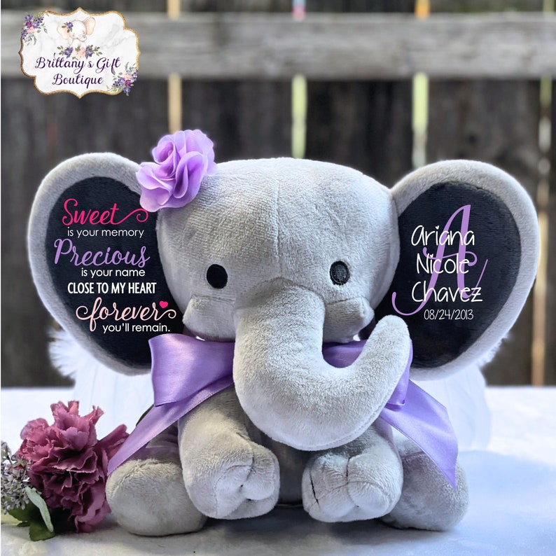 May include: A gray plush elephant with a purple bow and flower. The elephant has black eyes and a personalized message on its ear that reads "Ariana Nicole Chavez 08/24/2013". The other ear has the text "Sweet is your memory, Precious is your name, Close to my heart forever you'll remain."
