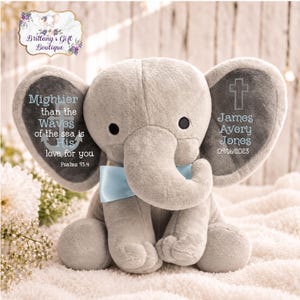 May include: A plush gray elephant toy featuring a light blue satin bow around its neck. The left ear displays an anchor icon and the text "Mightier than the Waves of the sea is His love for you Psalms 93:4". The right ear shows a cross and "James Avery Jones 09/16/2023".