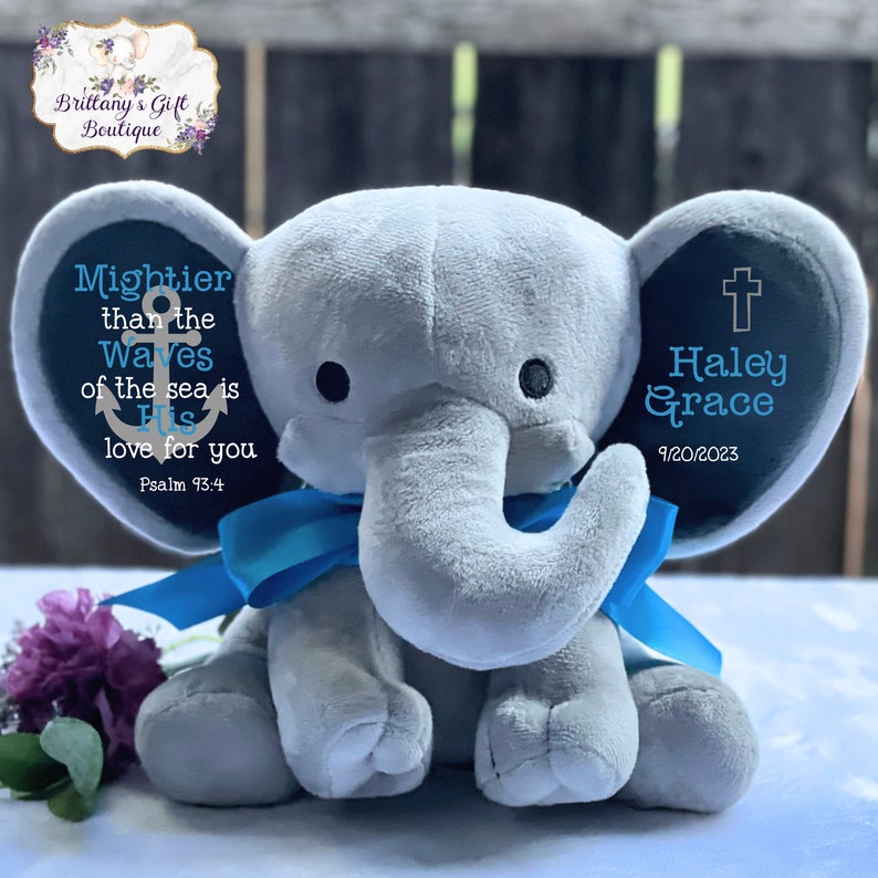 May include: A soft gray plush elephant toy featuring a bright blue ribbon tied around its neck. The elephant's ears are personalized with text: the left ear displays an anchor icon and the phrase Mightier than the Waves of the sea is His love for you Psalm 93:4, while the right ear shows a cross icon, the name Haley Grace, and the date 9/20/2023.