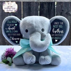 May include: A plush gray elephant with a light blue ribbon around its neck. The elephant's ears have text that reads "I'LL HOLD YOU IN MY heart" and "ONLY THE BEST BIG brothers ARE BROTHERS OF ANGELS". A purple flower and greenery are in the foreground.