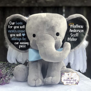 Może przedstawiać: Szary pluszowy słoń z niebieskimi skrzydłami i niebieską kokardką. Słoń ma czarne ucho z tekstem "Our Love for you will never cease you will always be our missing piece" i drugie czarne ucho z tekstem "Mathew Anderson Scott Miller".