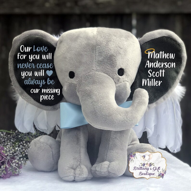 Może przedstawiać: Szary pluszowy słoń z niebieskimi skrzydłami i niebieską kokardką. Słoń ma czarne ucho z tekstem "Our Love for you will never cease you will always be our missing piece" i drugie czarne ucho z tekstem "Mathew Anderson Scott Miller".