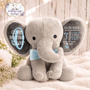May include: A plush gray elephant toy with a light blue bow tie. The left ear features the name Oliver Michael Taylor in blue and white text. The right ear displays birth details: 01-25-2024, 7 Pounds 10 Ounces, 21.5 inches, and 7:48 AM.