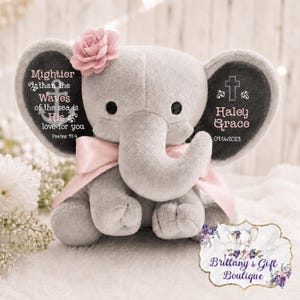 May include: A plush gray elephant toy featuring a pink fabric flower on its head and a pink ribbon around its neck. The left ear displays an anchor graphic with the text Mightier than the waves of the sea is His love for you Psalms 93:4.