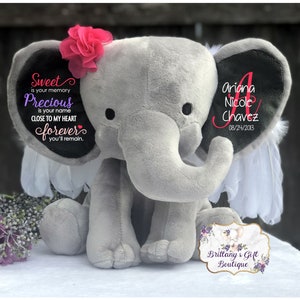 May include: A gray plush elephant with a pink flower on its head and white feather accents. The elephant's ear displays the name "Ariana Nicole Chavez" and the date "08/24/2013". Text on the other ear reads "Sweet is your memory, Precious is your name, Close to my heart forever, you'll remain."