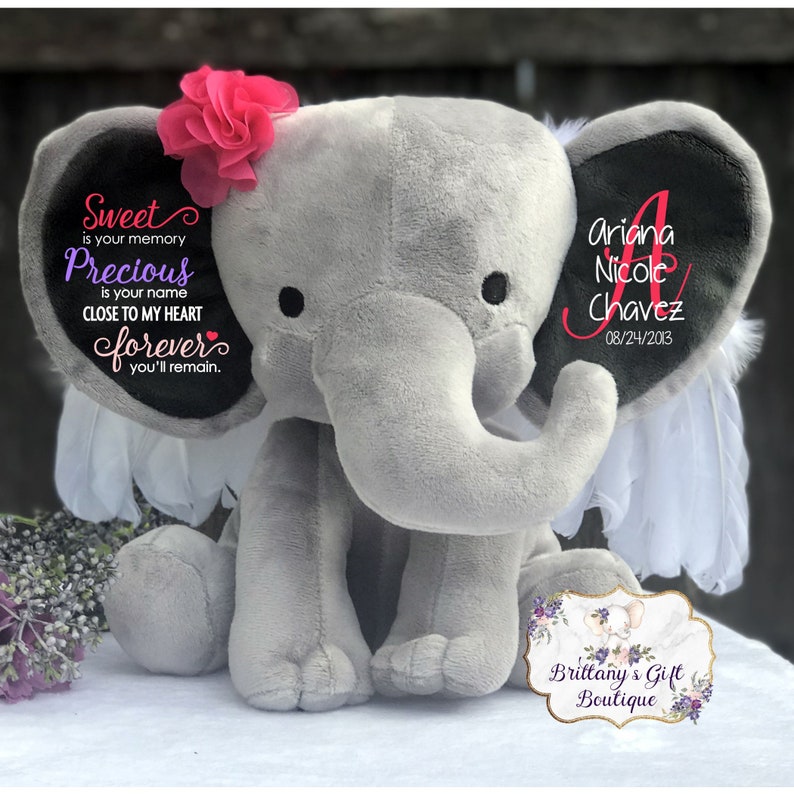 May include: A gray plush elephant with a pink flower on its head and white feather accents. The elephant's ear displays the name "Ariana Nicole Chavez" and the date "08/24/2013". Text on the other ear reads "Sweet is your memory, Precious is your name, Close to my heart forever, you'll remain."