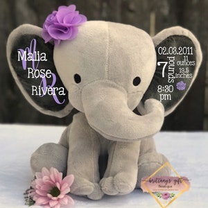 Puede incluir: Un peluche gris con forma de elefante con ojos negros, una flor morada y una flor rosa. Las orejas del elefante tienen el texto "Malia Rose Rivera" y detalles de nacimiento: 3,18 kg, 49,5 cm, y 20:30.