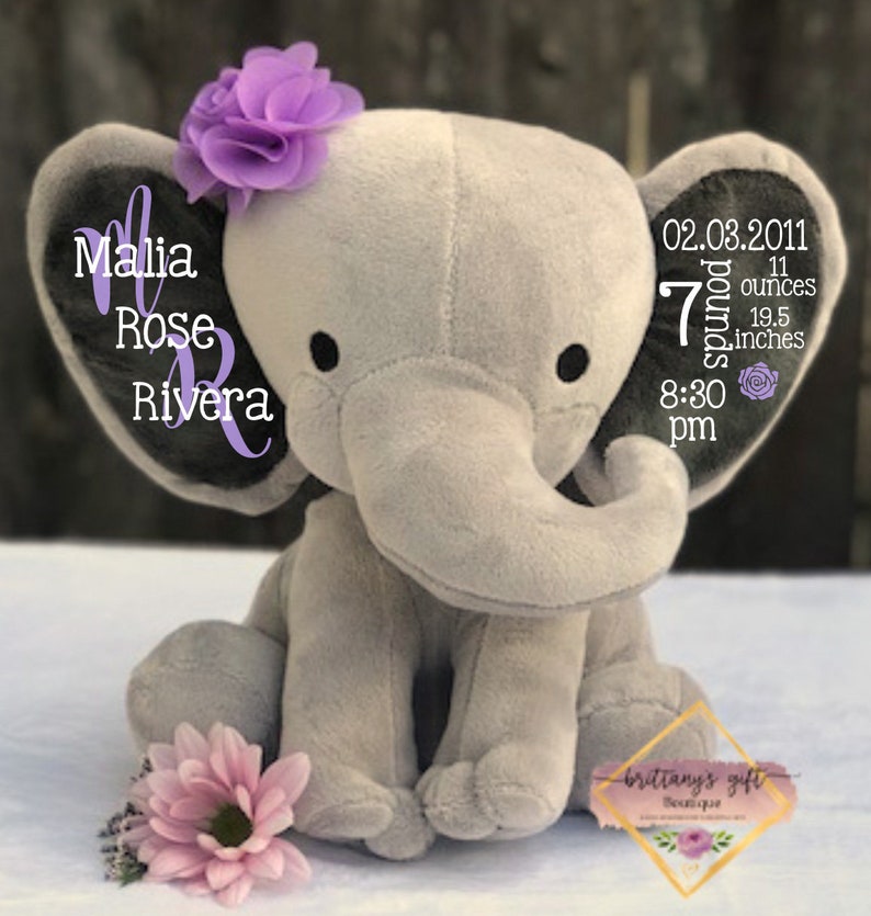 Puede incluir: Un peluche gris con forma de elefante con ojos negros, una flor morada y una flor rosa. Las orejas del elefante tienen el texto "Malia Rose Rivera" y detalles de nacimiento: 3,18 kg, 49,5 cm, y 20:30.