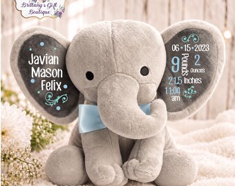 May include: A gray plush elephant toy with a light blue satin bow tie. The left ear features the name Javian Mason Felix in white and blue text. The right ear displays birth details: 06-15-2023, 9 Pounds 2 Ounces, 21.5 Inches, and 11:04 am.