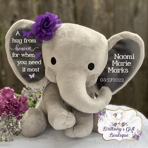 Puede incluir: Un elefante de peluche gris con ojos negros y una flor morada en la cabeza. Las orejas del elefante tienen el texto "A hug from heaven for when you need it most" y "Naomi Marie Marks 03/27/2022". Un regalo de Brittany's Gift Boutique.