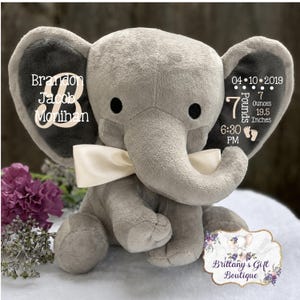 May include: A gray plush elephant toy with a cream-colored satin bow. The left ear features the text Brandon Jacob Monihan. The right ear displays the date 04-10-2019, 7 pounds 7 ounces, 19.5 inches, 6:30 PM, and two small footprint icons.