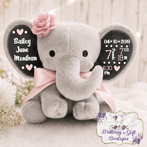 May include: A grey plush elephant toy featuring a pink fabric flower on its head and a pink satin ribbon around its neck. The ears display white text including the name Bailey Jane Monihan, the date 04-10-2019, and birth stats of 7 pounds 7 oz and 19 in.