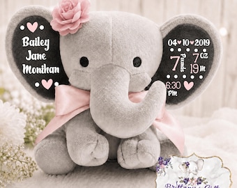 May include: A grey plush elephant toy featuring a pink fabric flower on its head and a pink satin ribbon around its neck. The ears display white text including the name Bailey Jane Monihan, the date 04-10-2019, and birth stats of 7 pounds 7 oz and 19 in.