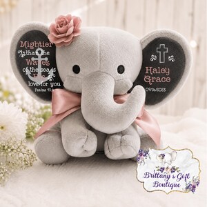 May include: A plush gray elephant toy featuring a pink fabric flower on its head and a pink ribbon around its neck. The ears display text including 'Mightier than the waves of the sea is His love for you Psalms 93:4', a cross, 'Haley Grace', and '09/16/2023'.