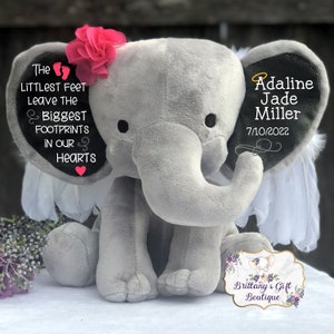 Op de afbeelding: Een grijze pluche olifant met witte vleugels en een roze bloem op zijn kop. De oren van de olifant zijn geborduurd met de tekst "The littlest feet leave the biggest footprints in our hearts" en "Adaline Jade Miller 7/10/2022".