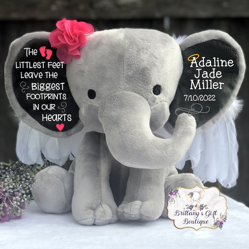 Op de afbeelding: Een grijze pluche olifant met witte vleugels en een roze bloem op zijn kop. De oren van de olifant zijn geborduurd met de tekst "The littlest feet leave the biggest footprints in our hearts" en "Adaline Jade Miller 7/10/2022".