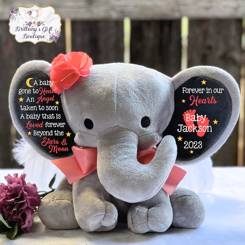 Puede incluir: Un elefante de peluche gris con una cinta rosa y una flor en la cabeza. Las orejas del elefante tienen parches de fieltro negro con el texto "A baby gone to Heaven, An Angel taken to soon, A baby that is Loved forever, Beyond the Stars & Moon" y "Forever in our Hearts, Baby Jackson, 2023".