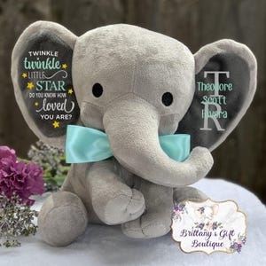 Puede incluir: Un peluche gris con forma de elefante y un lazo azul claro. Las orejas del elefante tienen el texto "Twinkle Twinkle Little Star" y un nombre. El juguete es de Brittany's Gift Boutique.