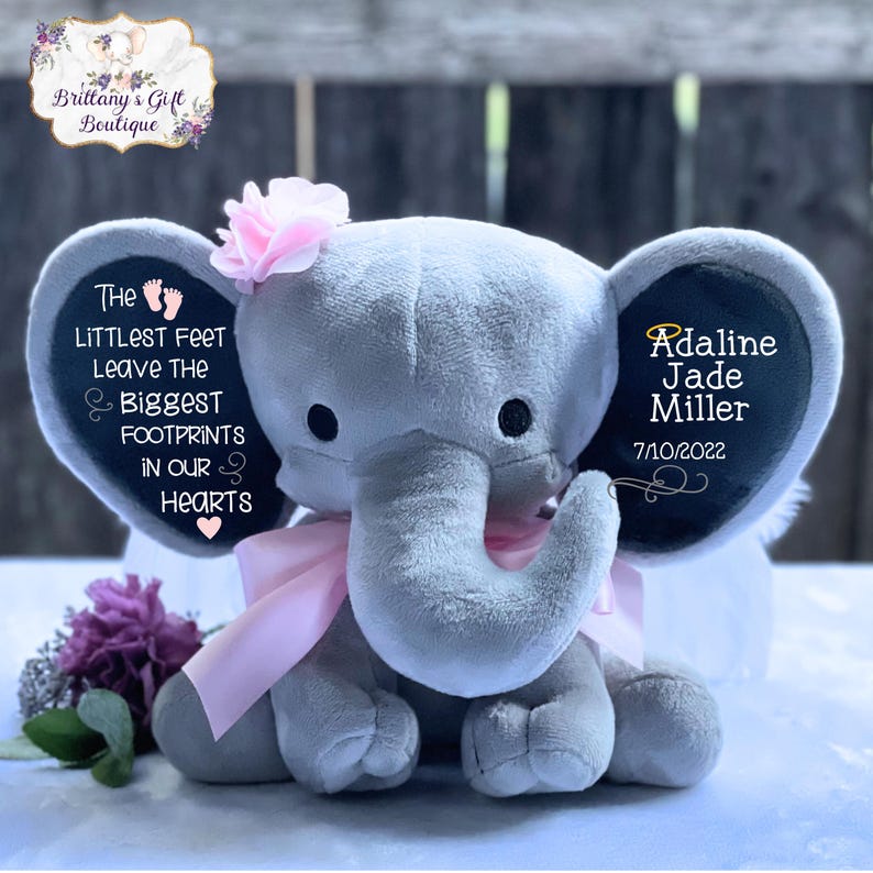 Op de afbeelding: Een grijze pluche olifant met een roze lint en een roze bloem op zijn kop. De olifant heeft een zwart vilt oor met de tekst "The littlest feet leave the biggest footprints in our hearts" en een ander zwart vilt oor met de tekst "Adaline Jade Miller 7/10/2022".
